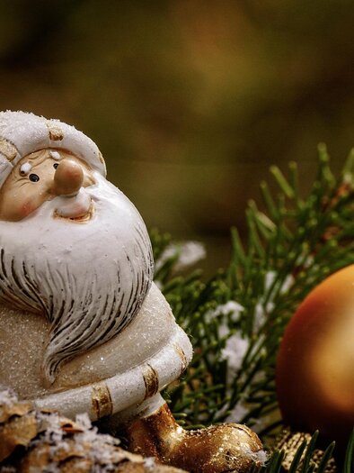 A small Christmas figure with a white beard and a red hat sits among pine branches. Next to her lies a golden Christmas ornament. | © pixabay