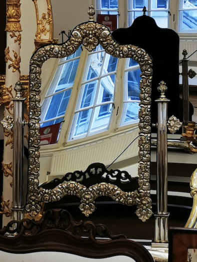 An elegant mirror with ornate decorations in silver and gold. In the background, more mirrors and windows can be seen. | © Irmgard-Unger-Eisele.