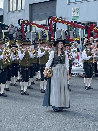 Eine Gruppe von Musikern in traditioneller Tracht zieht durch die Straße. Im Hintergrund sind Zuschauer versammelt, die das Ereignis beobachten. | © Musikverein Ratten