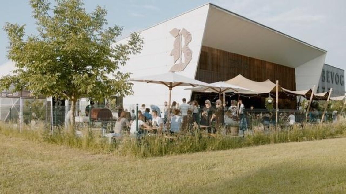 Ein modernes Café mit Außenbereich und vielen Gästen. Der Raum ist von einer grünen Wiese und Bäumen umgeben. | © Brauhaus Bevog