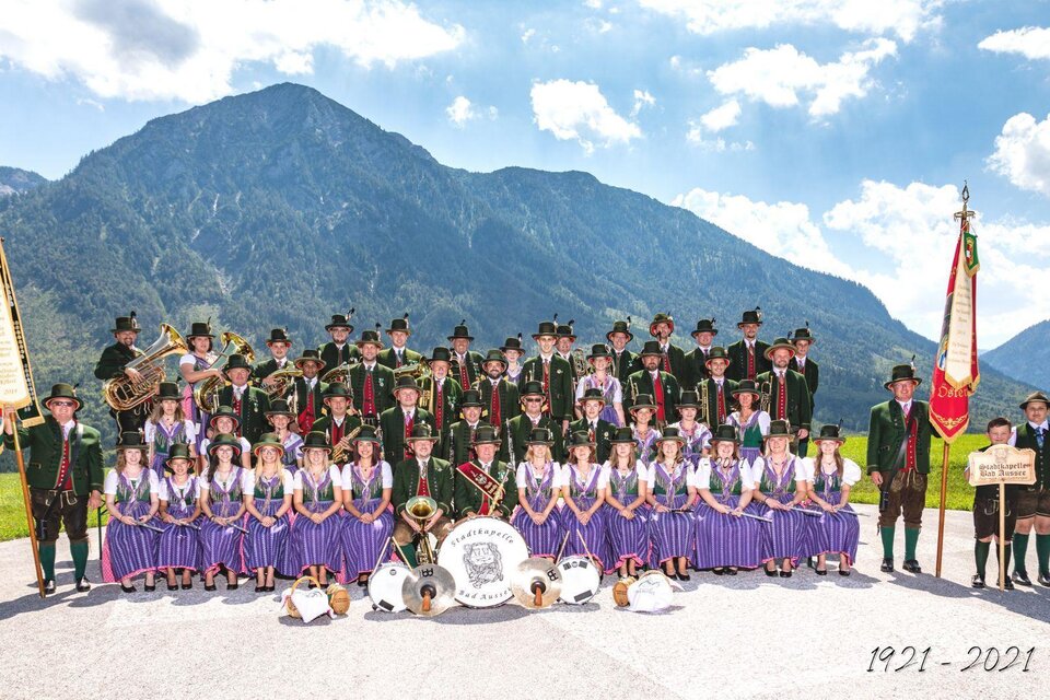 Eine große Gruppe von Menschen steht vor einer Berglandschaft. Sie tragen traditionelle Trachten und halten Musikinstrumente sowie Banner. | © Erich Kainzinger