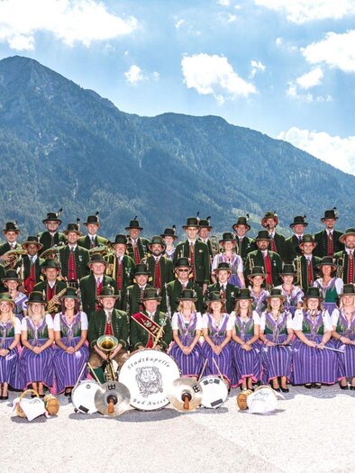 A large group of people stands in front of a mountain landscape. They are wearing traditional costumes and holding musical instruments as well as banners. | © Erich Kainzinger