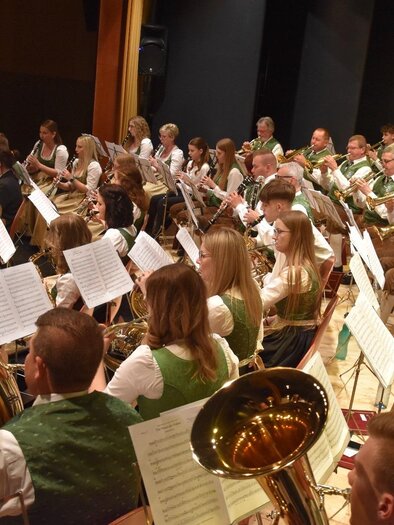 A large wind orchestra is playing on stage. The musicians are wearing traditional costumes and are focused on their sheet music. | © MMK Bad Waltersdorf