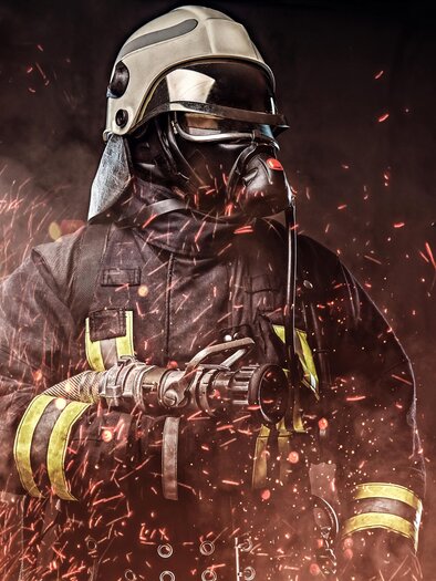 A firefighter in protective gear stands against a background of sparks and smoke. He wears a helmet and a breathing mask while holding a fire hose. | © px