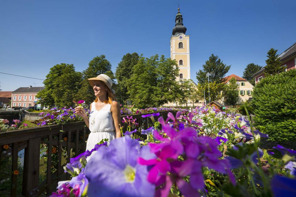 Eine Frau in einem weißen Kleid steht vor bunten Blumen und einem hohen Kirchturm. Der Himmel ist klar und blau, was eine fröhliche Atmosphäre schafft. | © Thermen-&Vulkanland_Bernhard Bergmann