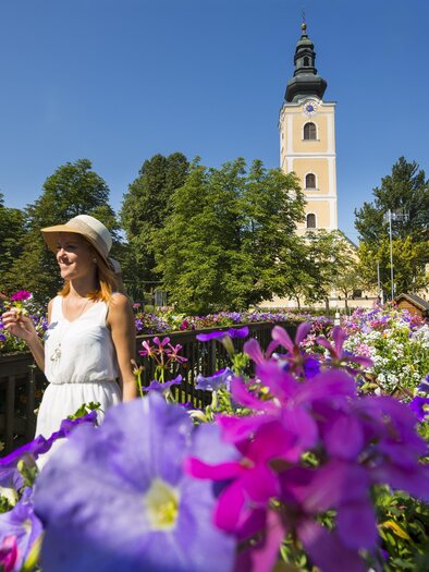 A woman in a white dress stands in front of colorful flowers and a tall church tower. The sky is clear and blue, creating a cheerful atmosphere. | © Thermen-&Vulkanland_Bernhard Bergmann