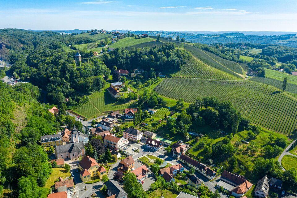 Eine malerische Landschaft mit sanften Hügeln und Weinbergen. Im Vordergrund sieht man ein kleines Dorf mit charmanten Häusern. | © TV Thermen- & Vulkanland