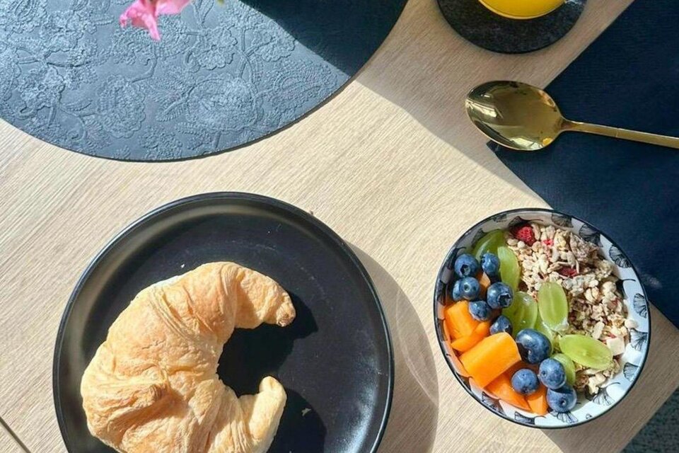 Ein leckerer Frühstückstisch mit einem Croissant und einer Schüssel Müsli, die mit frischen Früchten wie Heidelbeeren und Melonenstücken garniert ist. Im Hintergrund steht ein Glas Saft und eine Blume in einer Vase. | © Gipfelmomente GmbH