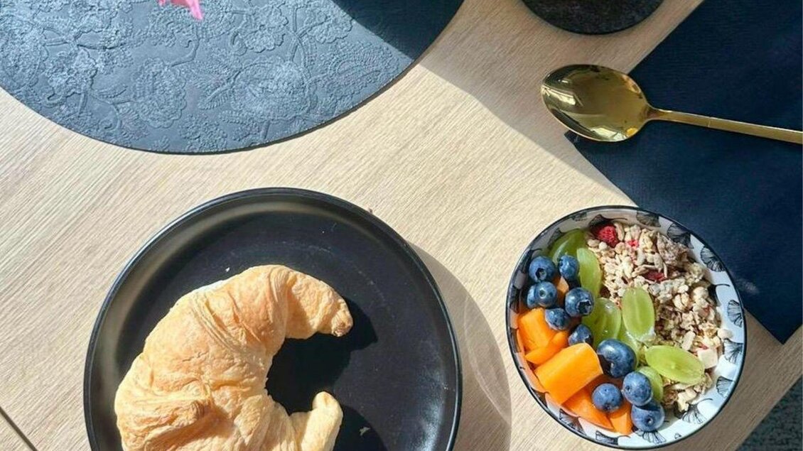 Ein leckerer Frühstückstisch mit einem Croissant und einer Schüssel Müsli, die mit frischen Früchten wie Heidelbeeren und Melonenstücken garniert ist. Im Hintergrund steht ein Glas Saft und eine Blume in einer Vase. | © Gipfelmomente GmbH