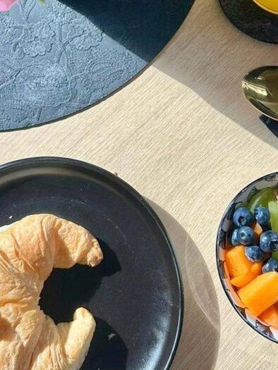 A delicious breakfast table with a croissant and a bowl of muesli garnished with fresh fruits like blueberries and pieces of melon. In the background, there is a glass of juice and a flower in a vase. | © Gipfelmomente GmbH