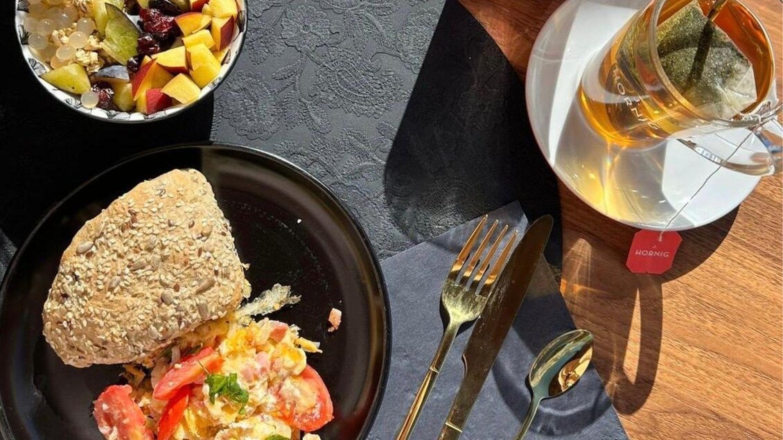 Ein Teller mit Rührei, Tomaten und einem Brötchen, daneben eine Schale mit Obst und ein Glas Tee. Die Szene ist gut beleuchtet und ansprechend angerichtet. | © Gipfelmomente GmbH