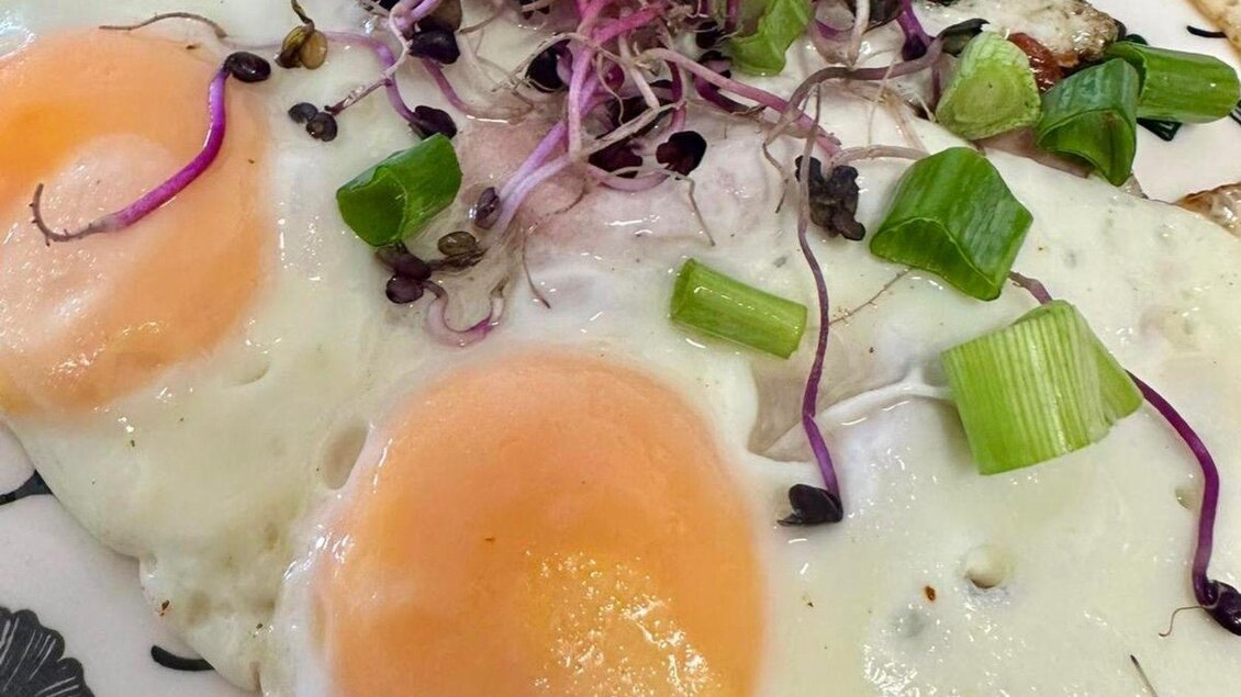 Zwei Spiegeleier mit orangefarbenem Eigelb, garniert mit frischen grünen Zwiebeln und Microgreens. Das Gericht ist auf einem dekorativen Teller angerichtet. | © Gipfelmomente GmbH