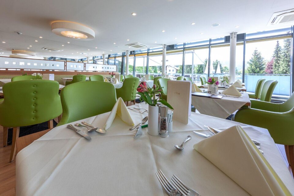 Ein modernes Restaurant mit großen Fenstern und hellen, grünen Stühlen. Die Tische sind elegant gedeckt mit weißen Tischdecken und frischen Blumen.