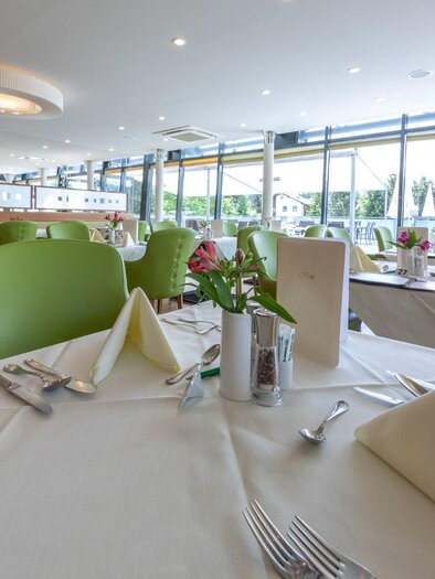 A modern restaurant with large windows and bright, green chairs. The tables are elegantly set with white tablecloths and fresh flowers.
