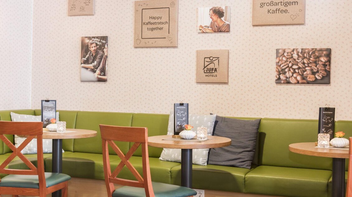 Ein gemütliches Café mit grünen Sitzbänken und Holzstühlen. An den Wänden hängen Bilder und Tafeln mit Kaffeethemen. | © JUFA Hotel Grundlsee