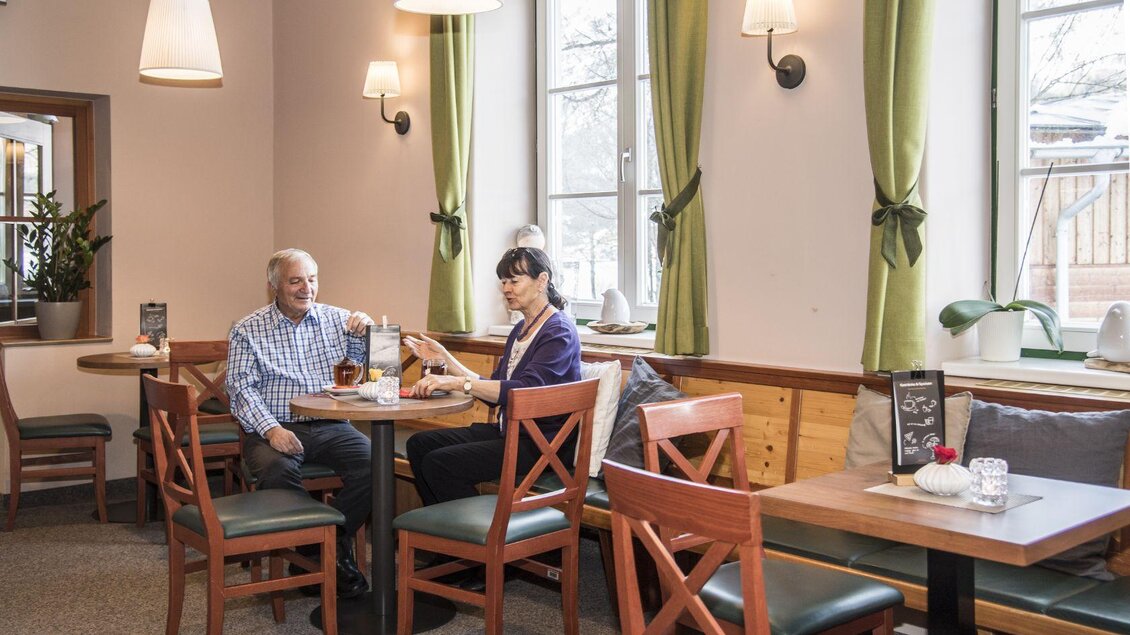 Ein gemütliches Café mit holzernen Tischen und Stühlen. Zwei Personen sitzen zusammen und genießen ihre Zeit bei einer Tasse Kaffee. | © JUFA Hotel Grundlsee