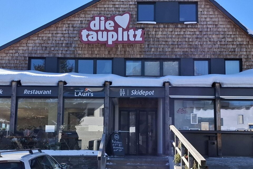 Ein gemütliches Restaurant mit einer großen Fensterfront und einem Schild mit dem Namen „die tauplitz“. Vor dem Gebäude liegt Schnee, und es sind einige Gäste zu sehen. | © TVB Ausseerland Salzkammergut/Raunigg