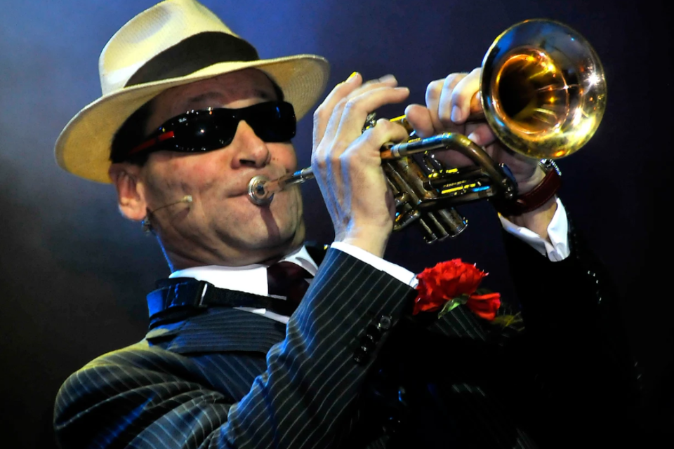 Ein Musiker spielt Trompete und trägt einen eleganten Anzug mit Hut und Sonnenbrille. Im Hintergrund ist eine atmosphärische Bühnenszene zu sehen. | © ©Scheuermann