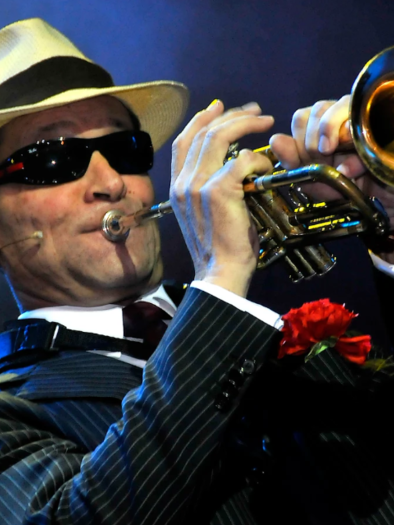 A musician plays the trumpet and wears an elegant suit with a hat and sunglasses. In the background, there is an atmospheric stage scene. | © ©Scheuermann