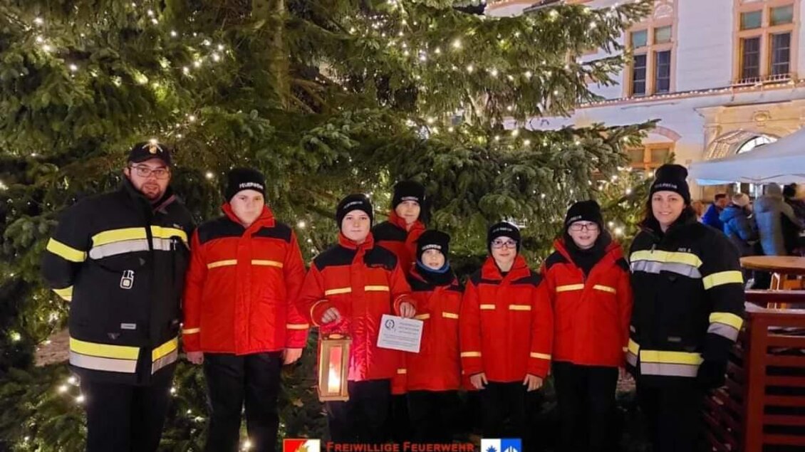 Eine Gruppe von Feuerwehrleuten in roten Jacken steht vor einem festlich geschmückten Weihnachtsbaum. Sie halten eine Laterne und ein Schild in der Hand.
