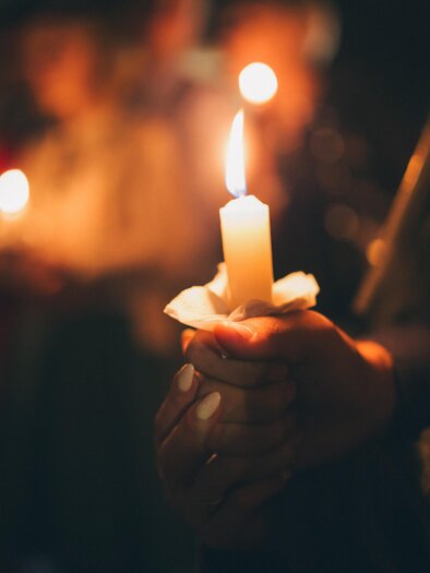 A person is holding a lit candle in their hand. In the background, other people are seen with candles. | © Adobe Stock