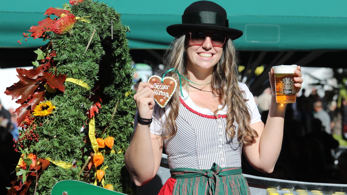 Eine fröhliche Frau trägt ein traditionelles Outfit und hält ein Bier sowie ein Lebkuchenherz in der Hand. Im Hintergrund sind festliche Dekorationen und Getränke sichtbar. | © Freisinger