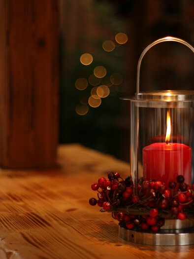 Ein stilvoller Laternen mit einer roten Kerze, umgeben von grünen Zweigen und Beeren, steht auf einem Holztisch. Im Hintergrund sind sanfte Lichter erkennbar, die eine gemütliche Atmosphäre schaffen. | © Tv Murau