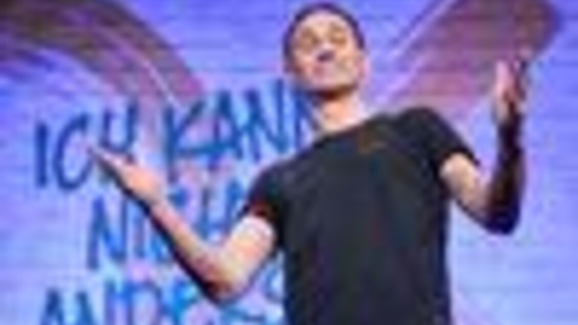 Ein Mann steht auf der Bühne und spricht interessiert sein Publikum an. Im Hintergrund ist ein lebendiges Wandbild mit dem Text „Ich kann nichts anderes“ zu sehen. | © ©MarkusWache