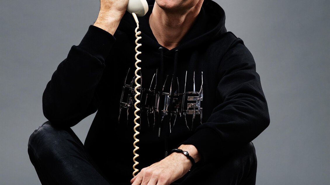 Ein Mann sitzt am Boden und hält einen alten Telefonhörer in der Hand. Er lächelt fröhlich und trägt einen schwarzen Hoodie. | © (c)-hans-leitner-klein
