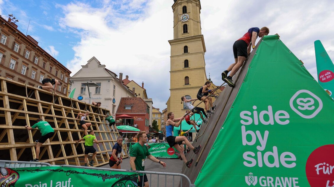 Eine sportliche Veranstaltung mit Teilnehmern, die an Kletterwänden arbeiten. Im Hintergrund steht ein historischer Turm.