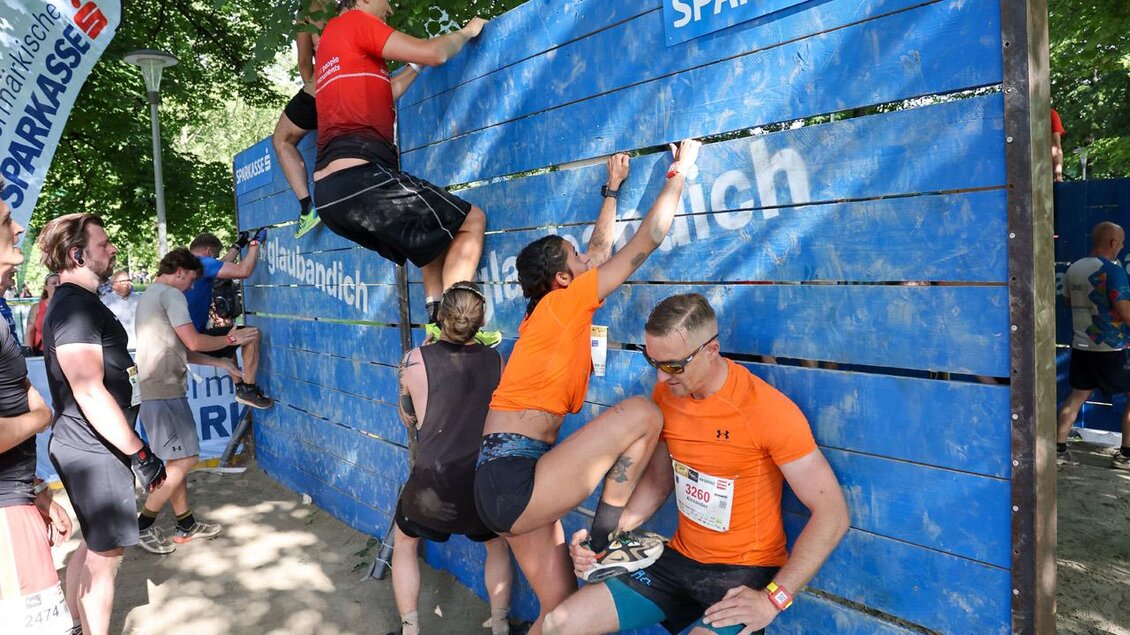 Eine Gruppe von Teilnehmern überwindet ein Hindernis bei einem Wettkampf. Es handelt sich um eine blaue Wand, die von mehreren Personen erklommen wird.