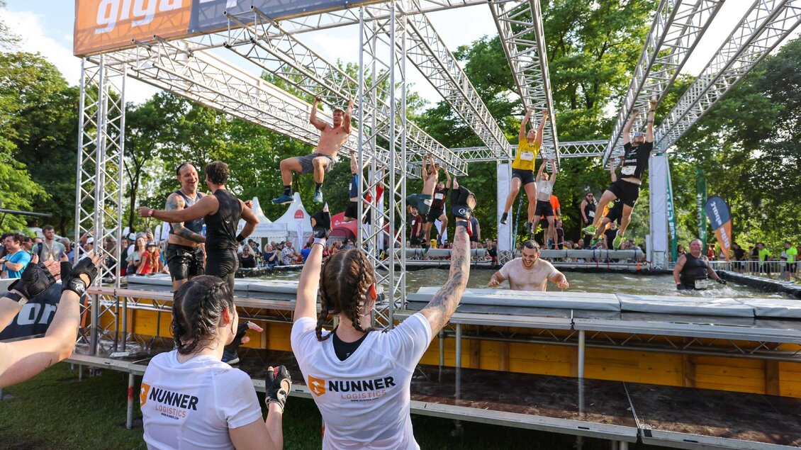 Eine Gruppe von Teilnehmern bewältigt einen Hindernislauf in einem Wasserspaßbereich. Zuschauer mit T-Shirts feuern die Sportler an.