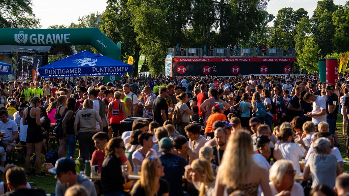 Eine große Menschenmenge versammelt sich im Freien bei einem Festival. Stände und Sponsorenbanner sind im Hintergrund zu sehen.