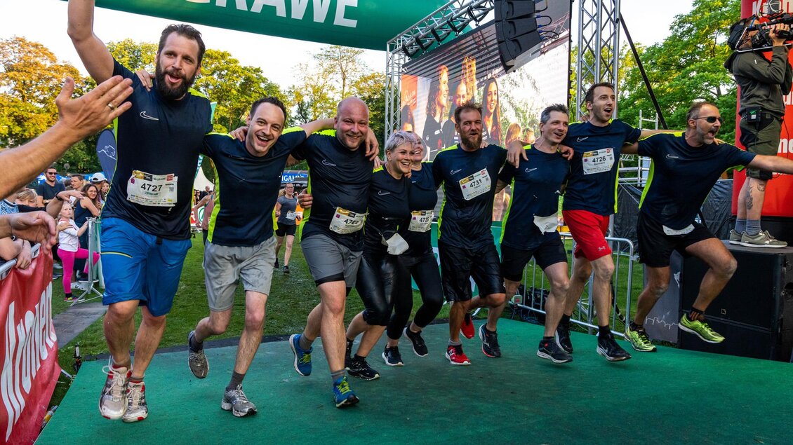 Eine Gruppe von Läufern feiert ihren Zieleinlauf bei einem Wettkampf. Sie tragen Sportkleidung und lächeln glücklich in die Kameras.