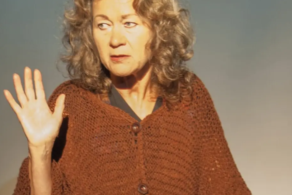 Eine ältere Frau mit lockigem, grauem Haar trägt einen braunen Poncho und schaut aufmerksam in die Kamera. Sie hebt ihre Hand, als ob sie etwas sagen oder zeigen möchte. | © KK