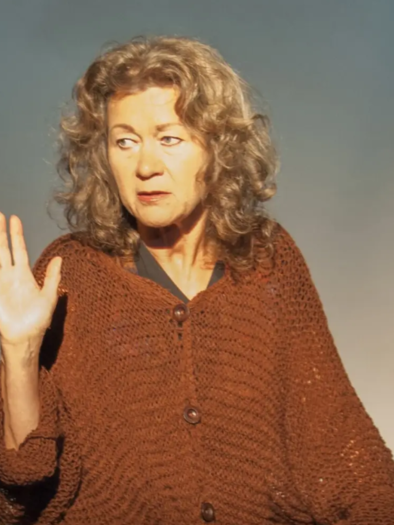 An older woman with curly gray hair is wearing a brown poncho and is looking attentively at the camera. She is raising her hand as if she wants to say or show something. | © KK
