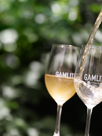 Two glasses with the print "GAMLIT," into which a drink is poured. In the background, green leaves are visible. | © IG Gamlitz / Stiefkind Fotografie