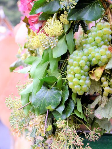 A decorative arrangement of grapes and plants hanging from a cart. In the background, other figures and a carriage are blurred. | © IG Tourismus Gamlitz