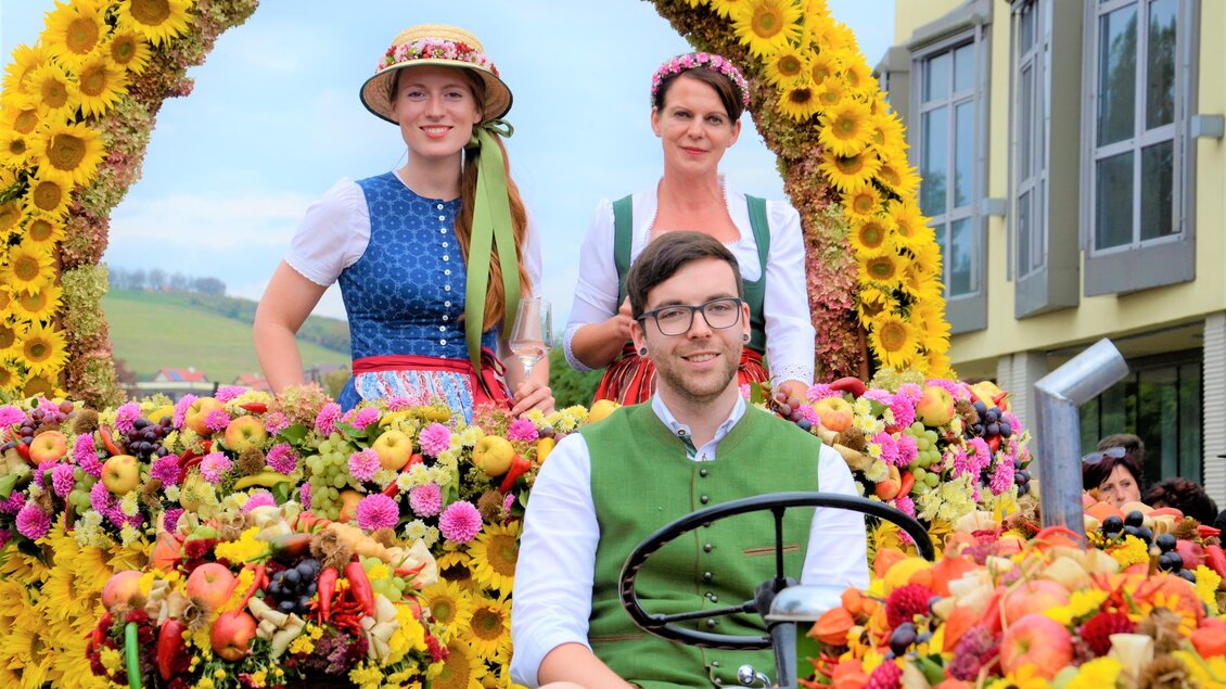 Ein festlicher Traktor mit bunten Blumen und fröhlichen Menschen. Die Frauen tragen traditionelle Trachten und lächeln in die Kamera. | © IG Tourismus Gamlitz