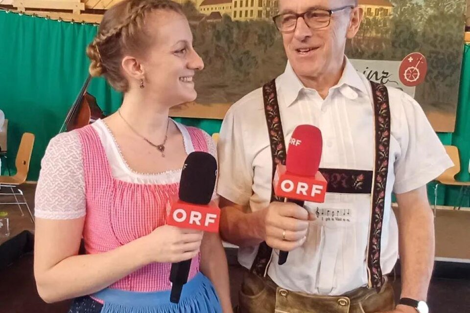 Ein Mann und eine Frau stehen in traditioneller Tracht mit Mikrofonen in der Hand. Sie sehen freundlich aus und scheinen ein Interview zu führen. | © Fam. Nöhrer
