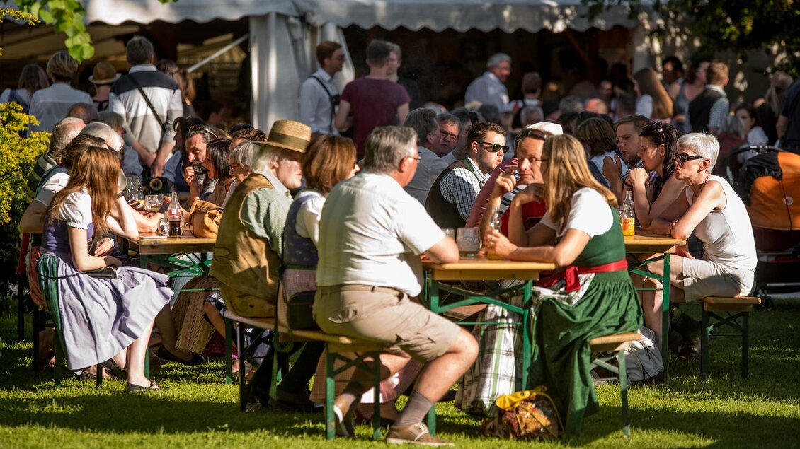 Eine Gruppe von Menschen sitzt an Tischen in einem Garten. Sie genießen das schöne Wetter und die Gesellschaft, während im Hintergrund weitere Besucher zu sehen sind. | © Narzissenfestverein/St. Pelizzari