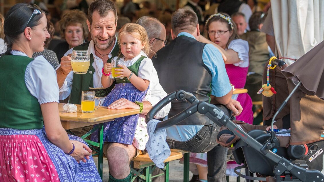 Eine fröhliche Szene auf einem Fest mit Familien am Tisch. Ein Mann hält ein Getränk, während ein kleines Mädchen an seiner Seite sitzt und ebenfalls etwas in der Hand hat. | © Narzissenfestverein/St. Pelizzari