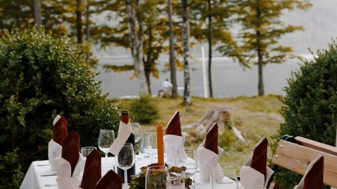 Ein schön gedeckter Tisch im Freien, umgeben von Bäumen und einer ruhigen Wasseroberfläche. Die Atmosphäre vermittelt eine gemütliche und einladende Stimmung. | © Prommer