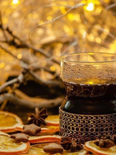 Ein Glas mit heißem Getränk steht auf einem Tisch, umgeben von getrockneten Orangenscheiben und Gewürzen. Im Hintergrund funkeln Lichter, die eine gemütliche Stimmung erzeugen. | © Pixabay