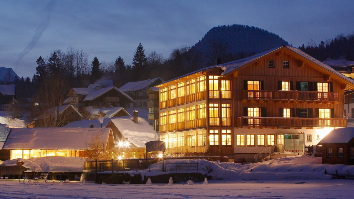 Ein schönes Gebäude im Schnee erleuchtet von warmem Licht. Im Hintergrund sind bewaldete Hügel und ein klarer Abendhimmel zu sehen. | © Seehotel Grundlsee