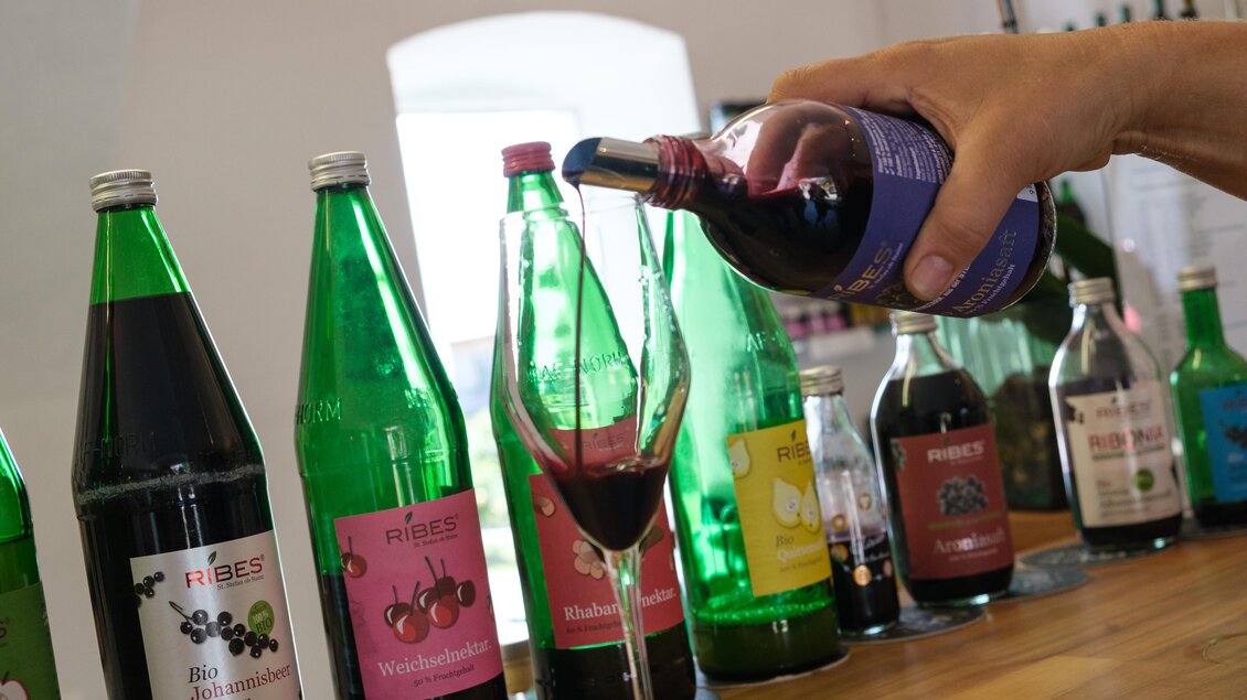 Ein Hand gießt ein Getränk aus einer Flasche in ein Glas. Im Hintergrund stehen verschiedene Flaschen mit fruchtigen Getränken. | © CDM