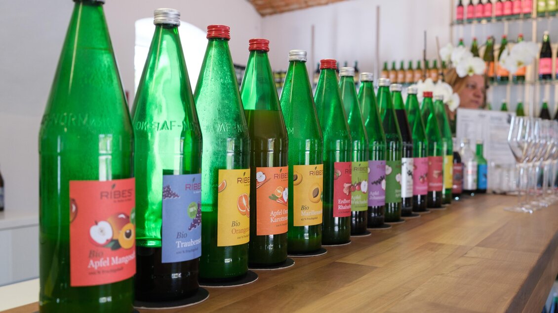 Eine Reihe grüner Flaschen mit bunten Etiketten steht auf einer Holztheke. Im Hintergrund sind weitere Flaschen und Gläser sichtbar. | © CDM