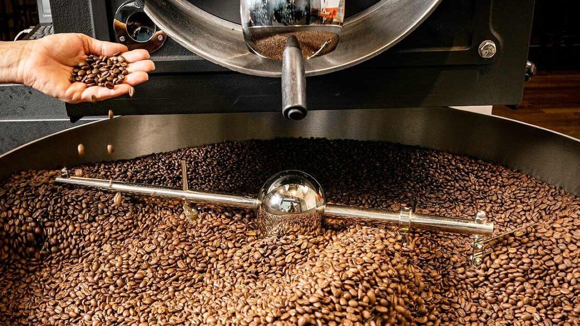 Eine handvoll Kaffeebohnen wird über einem großen Behälter mit gerösteten Bohnen gehalten. Der Behälter ist gefüllt mit einer Vielzahl von frisch gerösteten Kaffeebohnen. | © Cafe Hubmann