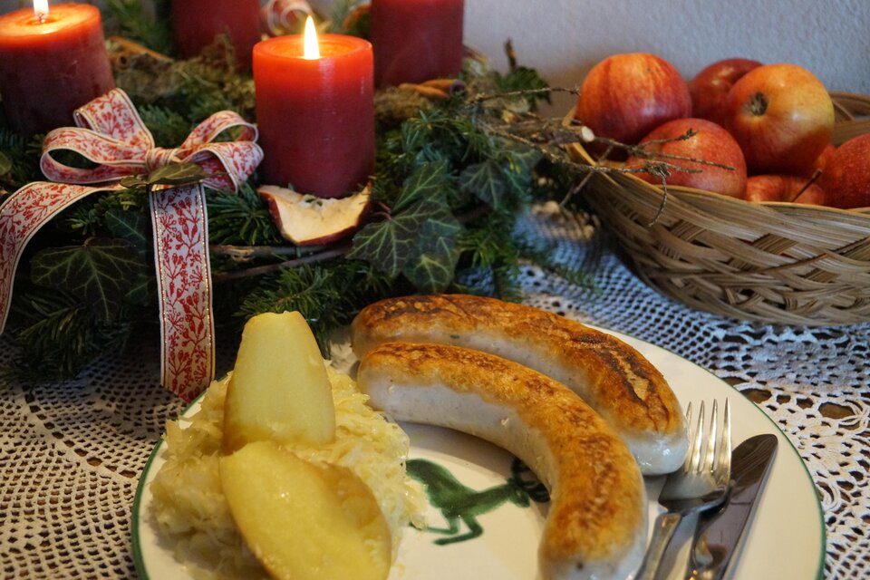 Ein festlich gedeckter Tisch mit Bratwürsten, Sauerkraut und Äpfeln. Im Hintergrund leuchten rote Kerzen auf einem Adventskranz. | © Marianne Ritzinger 