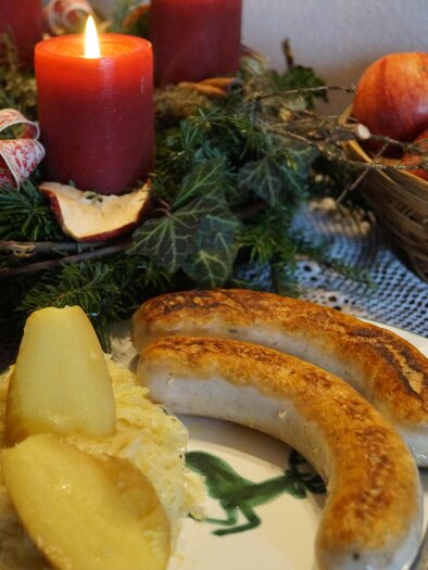 A festively set table with sausages, sauerkraut, and apples. In the background, red candles glow on an Advent wreath. | © Marianne Ritzinger 